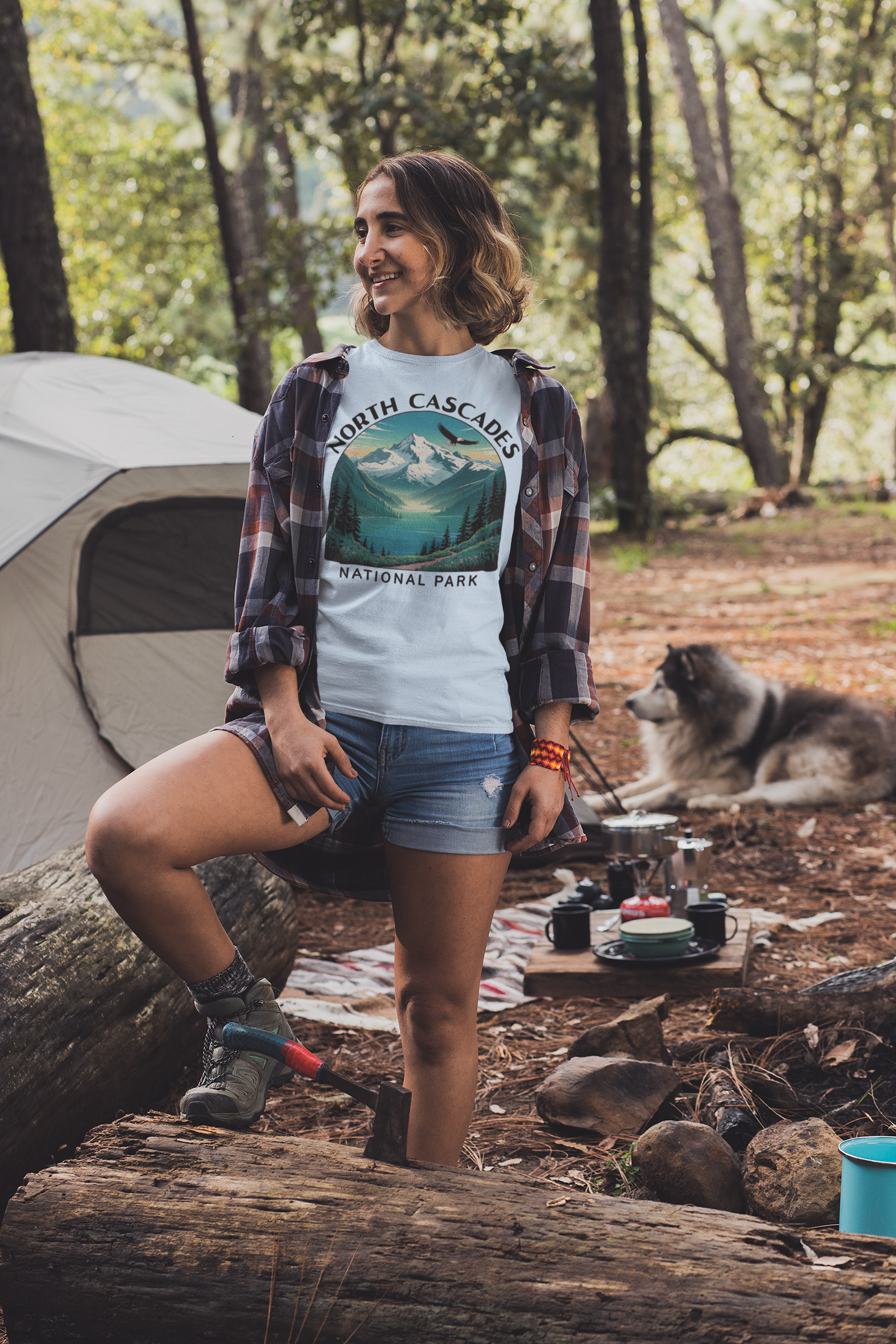 Woman in a National Park t-shirt sitting by a campsite with a dog and tent in the background.