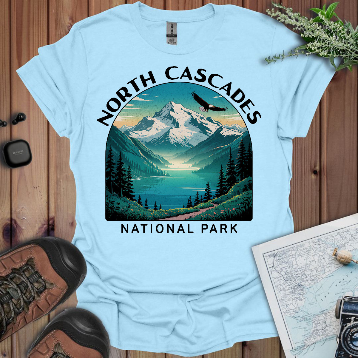 Light blue t-shirt with North Cascades National Park graphic on a wooden surface with hiking gear.