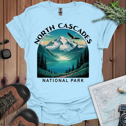 Light blue t-shirt with North Cascades National Park graphic on a wooden surface with hiking gear.