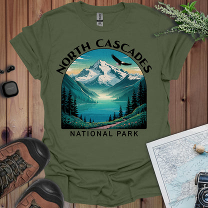 Green t-shirt with North Cascades National Park graphic on a wooden surface with hiking boots and map.