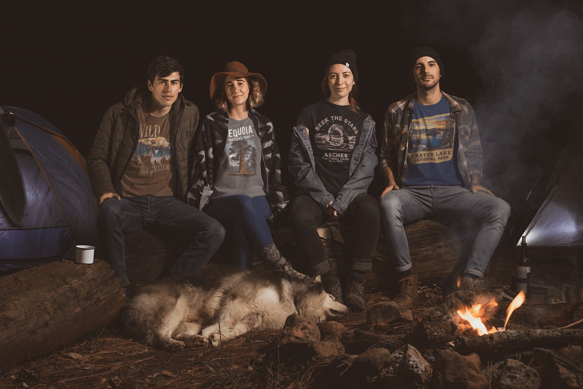 Four people and a dog sitting around a campfire at night with tents in the background.
